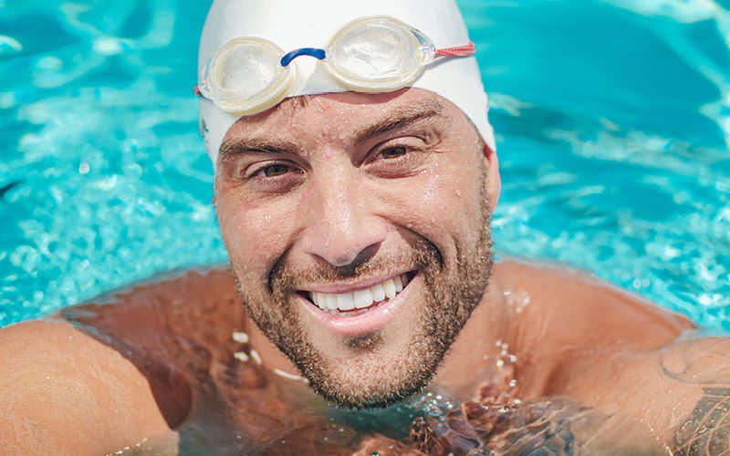 man in pool with cap and goggles, smiles at the camera